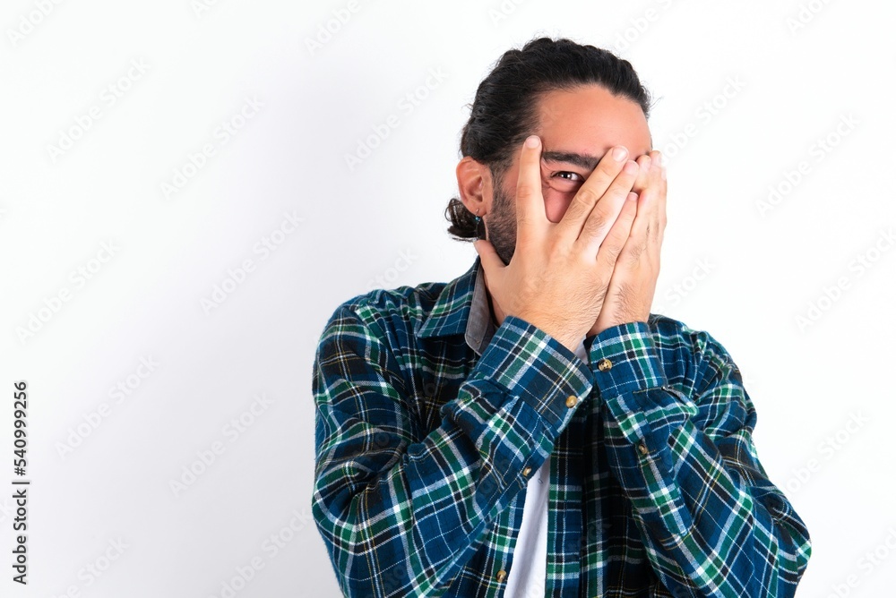 young bearded hispanic man wearing plaid shirt over white background covering face with hands and peering out with one eye between fingers. Scared from something or someone. young bearded hispanic man wearing plaid shirt over white background covering face with hands and peering out with one eye between fingers. Scared from something or someone.
