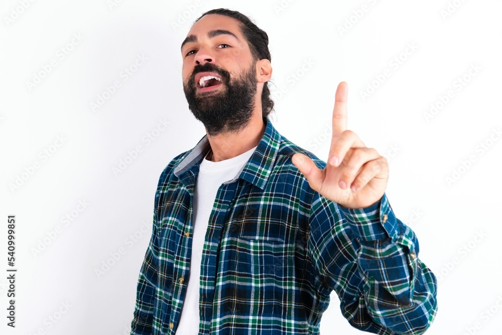 Pleasant looking young bearded hispanic man wearing plaid shirt over white background has clever expression, raises one finger, remembers himself not to forget tell important thing. Pleasant looking young bearded hispanic man wearing plaid shirt over white background has clever expression, raises one finger, remembers himself not to forget tell important thing.