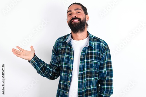 Positive glad young bearded hispanic man wearing plaid shirt over white says: wow how exciting it is, has amazed expression, shows something on blank space with open hand. Advertisement concept.