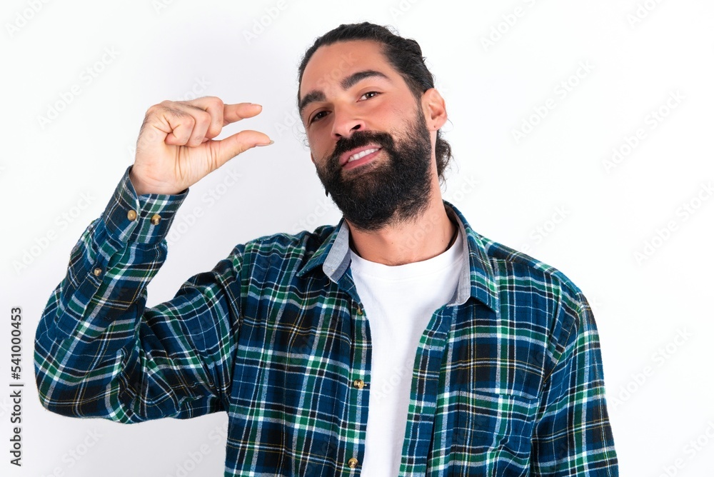 Fototapeta premium Shocked young bearded hispanic man wearing plaid shirt over white background shows something little with hands, demonstrates size, opens mouth from surprise. Measurement concept.