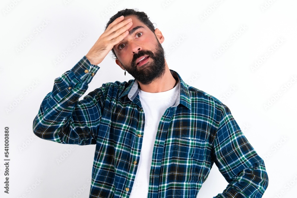 young bearded hispanic man wearing plaid shirt over white background touching forehead, hears something surprising, glad receive good news, feels relieved. Almost got in trouble. young bearded hispanic man wearing plaid shirt over white background touching forehead, hears something surprising, glad receive good news, feels relieved. Almost got in trouble.