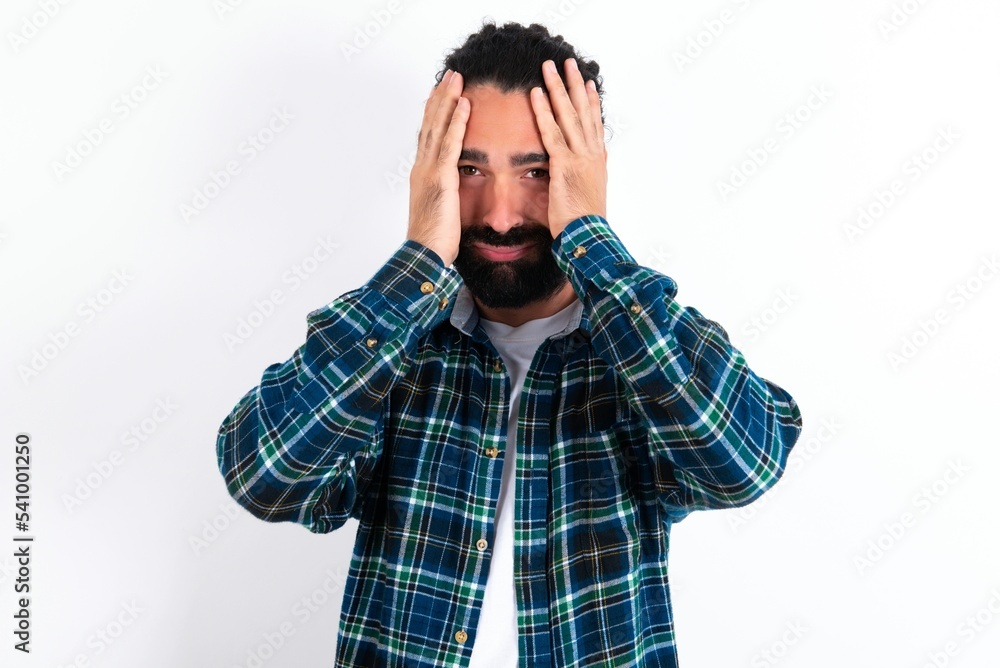 young bearded hispanic man wearing plaid shirt over white background holding head in hands with unhappy expression watching sad movie about animals and trying not to cry. young bearded hispanic man wearing plaid shirt over white background holding head in hands with unhappy expression watching sad movie about animals and trying not to cry.