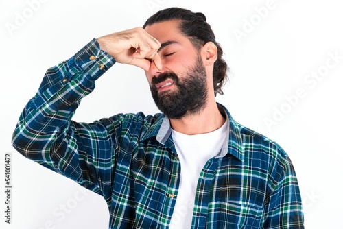 Displeased young bearded hispanic man wearing plaid shirt over white background plugs nose as smells something stink and unpleasant, feels aversion, hates disgusting scent.