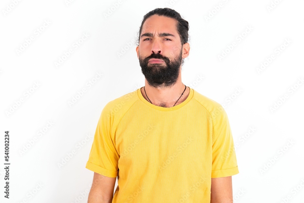 Offended dissatisfied young bearded hispanic man wearing yellow T-shirt over white background with moody displeased expression at camera being disappointed by something Offended dissatisfied young bearded hispanic man wearing yellow T-shirt over white background with moody displeased expression at camera being disappointed by something