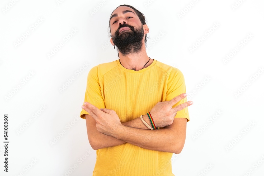 young bearded hispanic man wearing yellow T-shirt over white background crosses arms and points at different sides hesitates between two items or variants. Needs help with decision young bearded hispanic man wearing yellow T-shirt over white background crosses arms and points at different sides hesitates between two items or variants. Needs help with decision