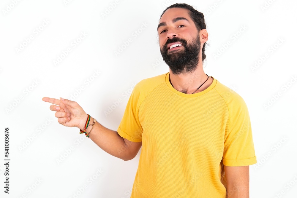 young bearded hispanic man wearing yellow T-shirt over white background laughs happily points away on blank space demonstrates shopping discount offer, excited by good news or unexpected sale. young bearded hispanic man wearing yellow T-shirt over white background laughs happily points away on blank space demonstrates shopping discount offer, excited by good news or unexpected sale.