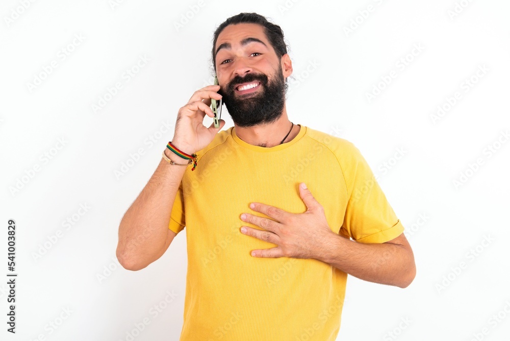 Smiling young bearded hispanic man wearing yellow T-shirt over white background talks via cellphone, enjoys pleasant great conversation. People, technology, communication concept Smiling young bearded hispanic man wearing yellow T-shirt over white background talks via cellphone, enjoys pleasant great conversation. People, technology, communication concept