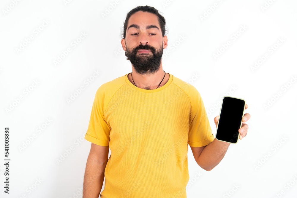 young bearded hispanic man wearing yellow T-shirt over white background holds new mobile phone and looks mysterious aside shows blank display of modern cellular young bearded hispanic man wearing yellow T-shirt over white background holds new mobile phone and looks mysterious aside shows blank display of modern cellular