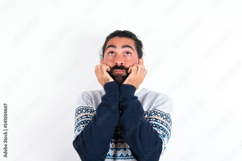 Portrait of sad young bearded hispanic man wearing knitted sweater over white background hands face look empty space Portrait of sad young bearded hispanic man wearing knitted sweater over white background hands face look empty space