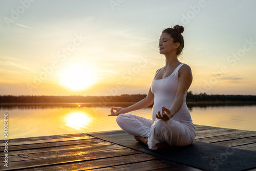 Pregnant woman doing yoga at lake during sunset.