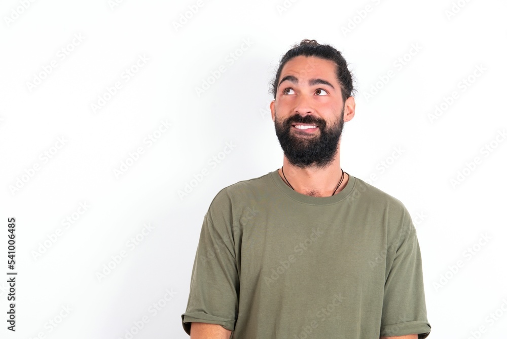 Fototapeta premium Amazed young bearded hispanic man wearing green T-shirt over white background bitting lip and looking tricky to empty space.
