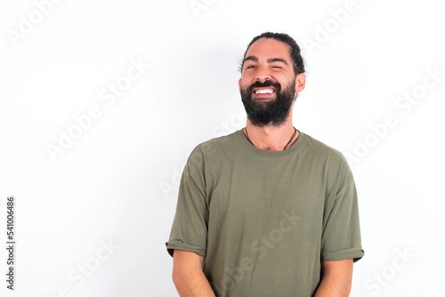 Coquettish young bearded hispanic man wearing green T-shirt over white background smiling happily, blinking at camera in a playful manner, flirting with you.