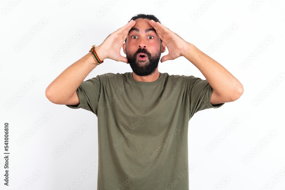 young bearded hispanic man wearing green T-shirt over white background with scared expression, keeps hands on head, jaw dropped, has terrific expression. Omg concept young bearded hispanic man wearing green T-shirt over white background with scared expression, keeps hands on head, jaw dropped, has terrific expression. Omg concept