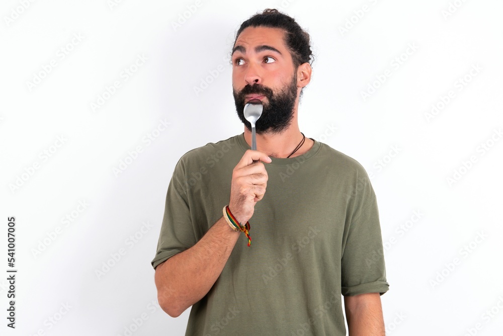 Very hungry young bearded hispanic man wearing green T-shirt over white background holding spoon into mouth dream of tasty meal