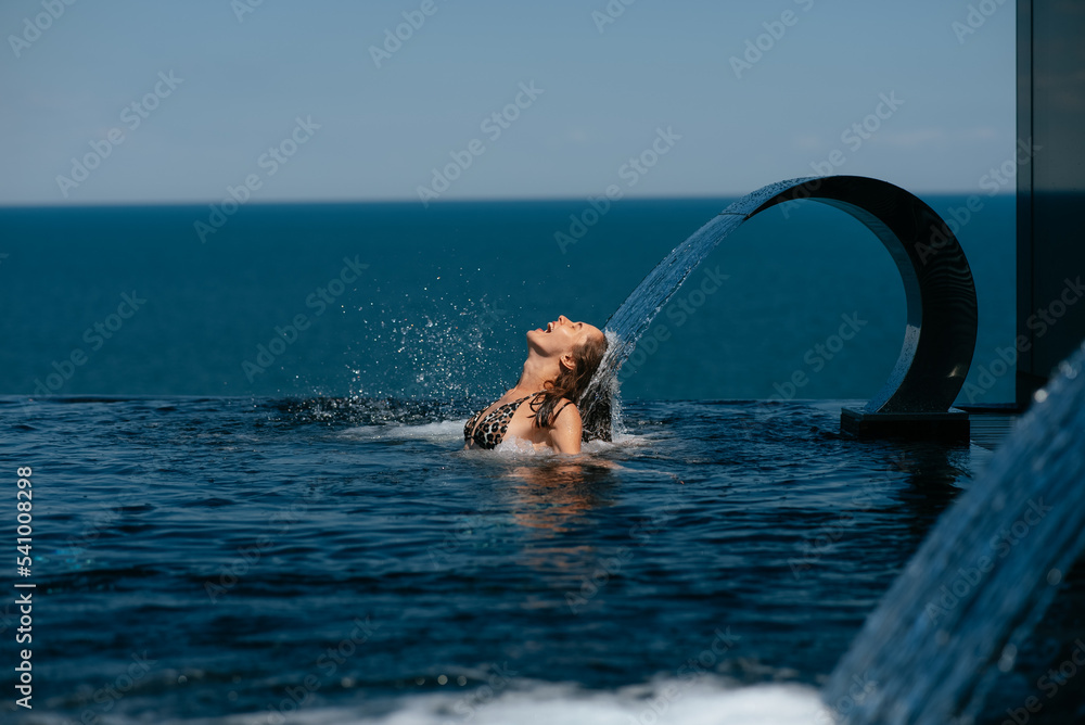 Fototapeta premium Young woman relaxing under swimming pool waterfall jet