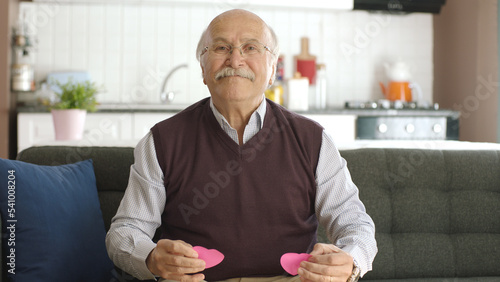 Happy old man looking at camera with two paper hearts in hands. Old man on the sofa of his house preparing to shoot a video of a message to his wife with a red paper heart. 