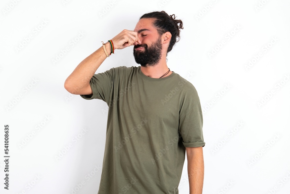 Fototapeta premium young bearded hispanic man wearing green t-shirt over white background smelling something stinky and disgusting, intolerable smell, holding breath with fingers on nose. Bad smell