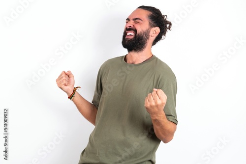 young bearded hispanic man wearing green t-shirt over white background celebrating surprised and amazed for success with arms raised and eyes closed. Winner concept. © Jihan