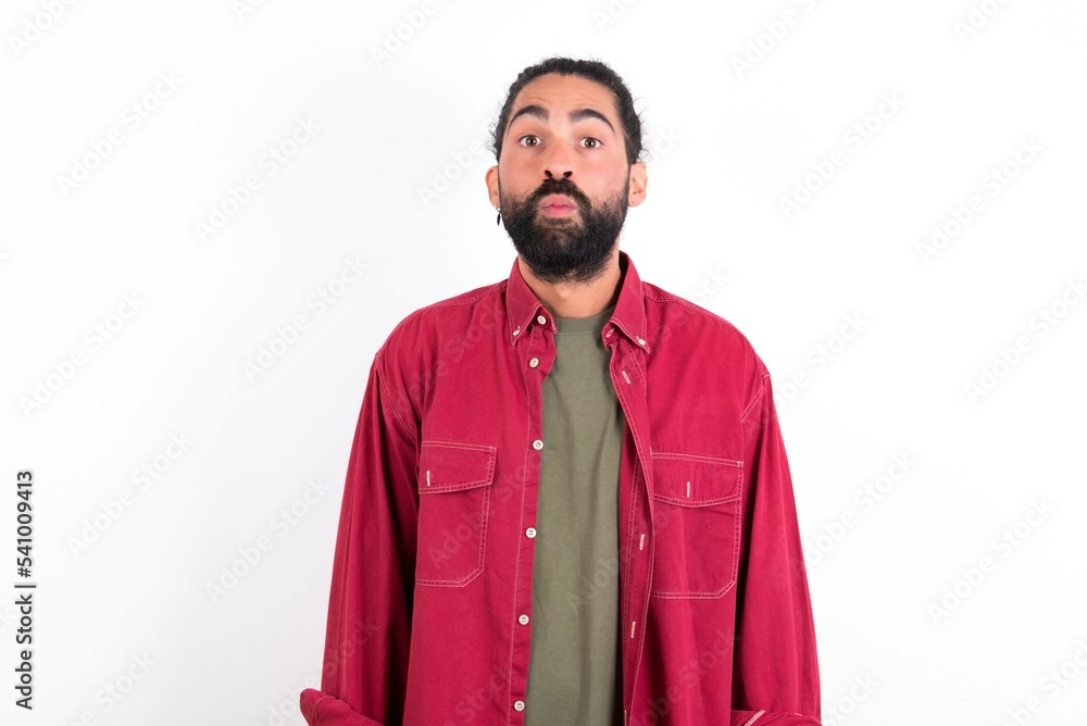Shot of pleasant looking young bearded hispanic man wearing red overshirt over white background, pouts lips, looks at camera, Human facial expressions Shot of pleasant looking young bearded hispanic man wearing red overshirt over white background, pouts lips, looks at camera, Human facial expressions