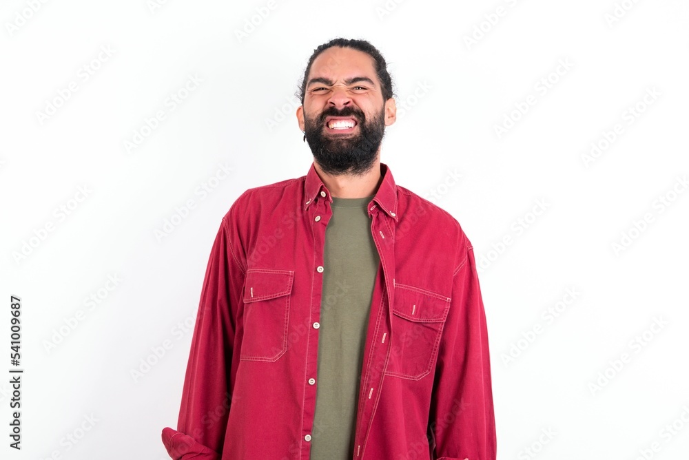 Caucasian man with beard wearing red shirt over white background keeps teeth clenched, frowns face in dissatisfaction, irritated because of much duties. Caucasian man with beard wearing red shirt over white background keeps teeth clenched, frowns face in dissatisfaction, irritated because of much duties.
