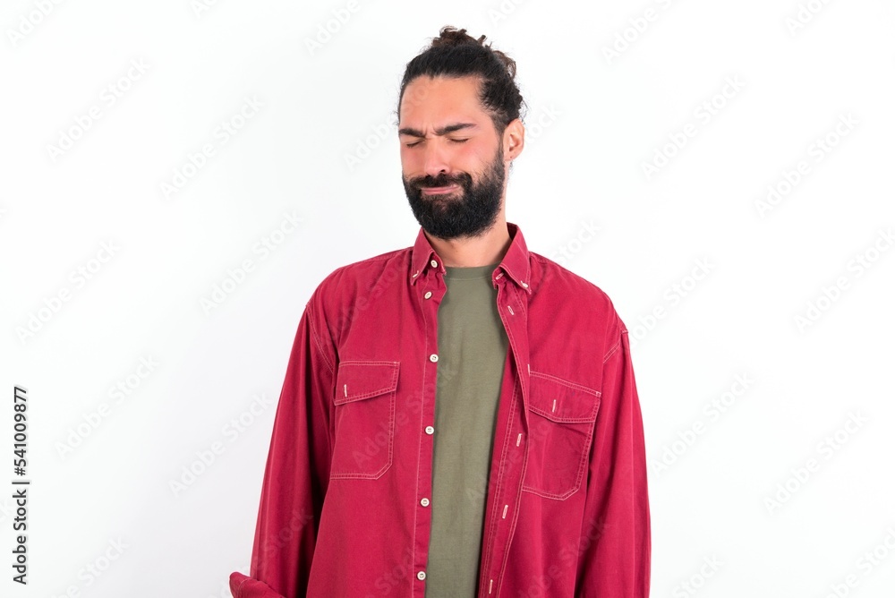Dismal gloomy rejected Caucasian man with beard wearing red shirt over white background has problems and difficulties, curves lower lip and closes eyes in despair, being in depression Dismal gloomy rejected Caucasian man with beard wearing red shirt over white background has problems and difficulties, curves lower lip and closes eyes in despair, being in depression