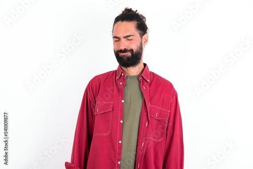 Dismal gloomy rejected Caucasian man with beard wearing red shirt over white background has problems and difficulties, curves lower lip and closes eyes in despair, being in depression