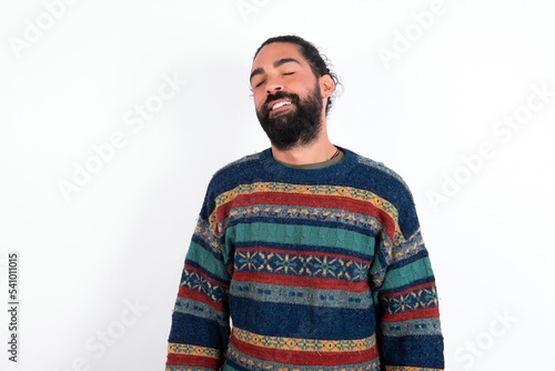 Positive Caucasian man with beard wearing sweater over white background with overjoyed expression closes eyes and laughs shows white perfect teeth