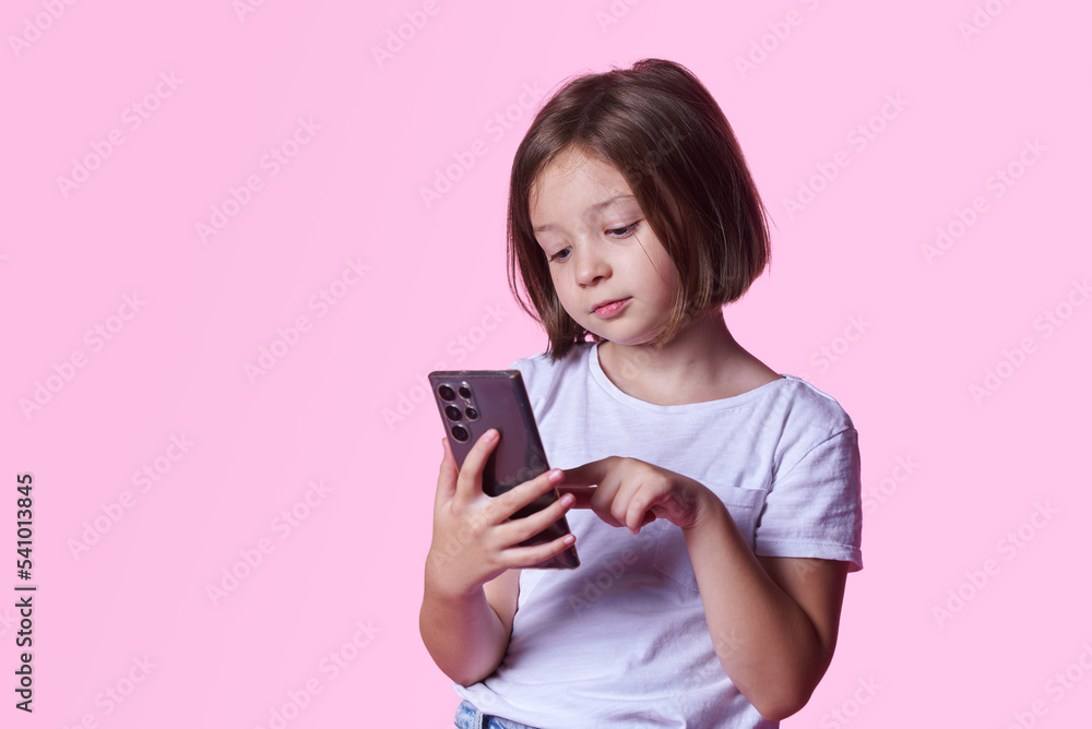 Addicted to social media: young preteen girl browsing internet on her big smartphone, pink studio background Addicted to social media: young preteen girl browsing internet on her big smartphone, pink studio background