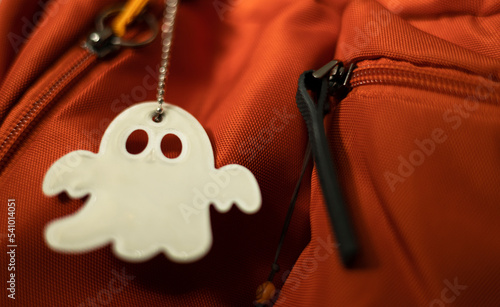 Key chain reflector in ghost pattern form on orange backpack, security sign to protect pedestrian visibility from vehicles on the road. Accessory essential for passerby to walk on the dark street