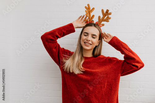 Lovely cute charm in lady with blond hair wearing red pullover and Christmas accessories on head is closed eyes and raised up hands on isolated background 