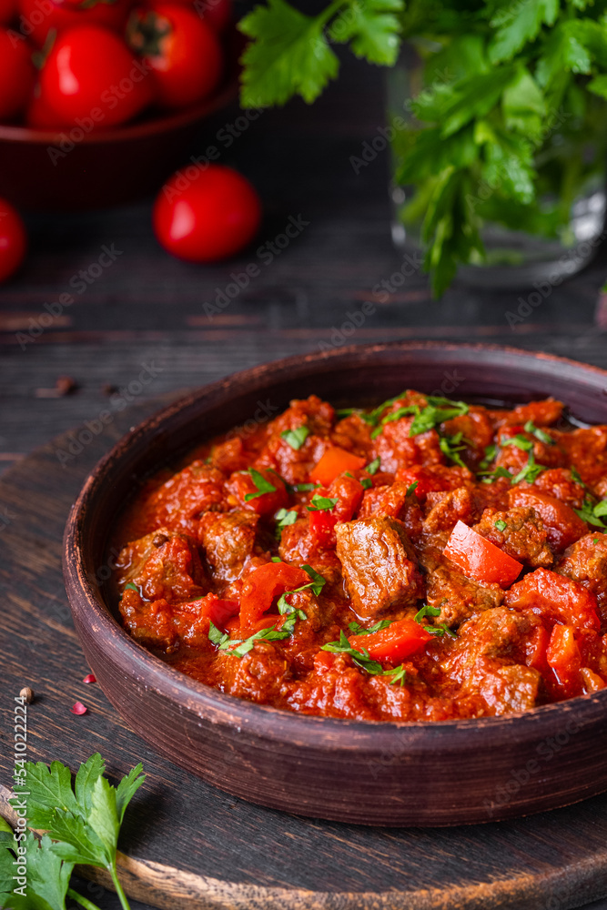 Chashushuli, thick beef stew in tomato sauce with sweet pepper, onion and spices in a clay bowl on a dark wooden background. Beef recipes. Chashushuli, thick beef stew in tomato sauce with sweet pepper, onion and spices in a clay bowl on a dark wooden background. Beef recipes.