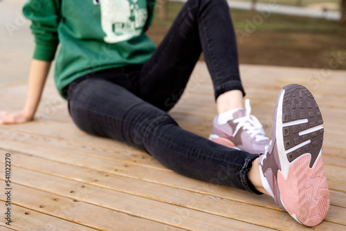 An unrecognizable woman in green sweater and black jeans sits on wooden deck. Womens legs in pink sneakers. Outsole of sports shoes. Side view