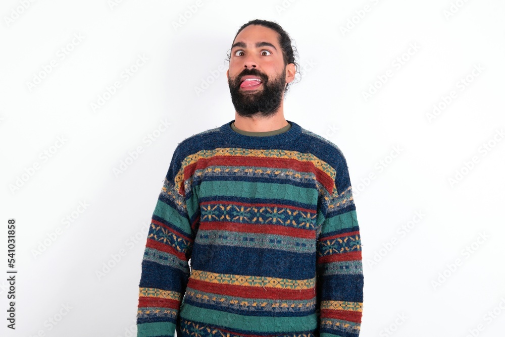 Fototapeta premium Funny Caucasian man with beard wearing sweater over white background makes grimace and crosses eyes plays fool has fun alone sticks out tongue.