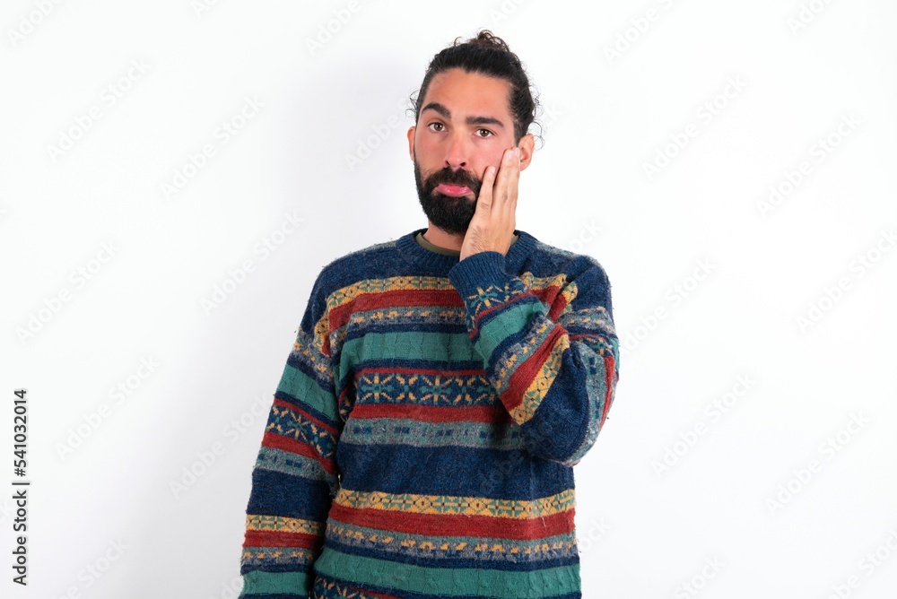 Sad lonely Caucasian man with beard wearing sweater over white background touches cheek with hand bites lower lip and gazes with displeasure. Bad emotions Sad lonely Caucasian man with beard wearing sweater over white background touches cheek with hand bites lower lip and gazes with displeasure. Bad emotions