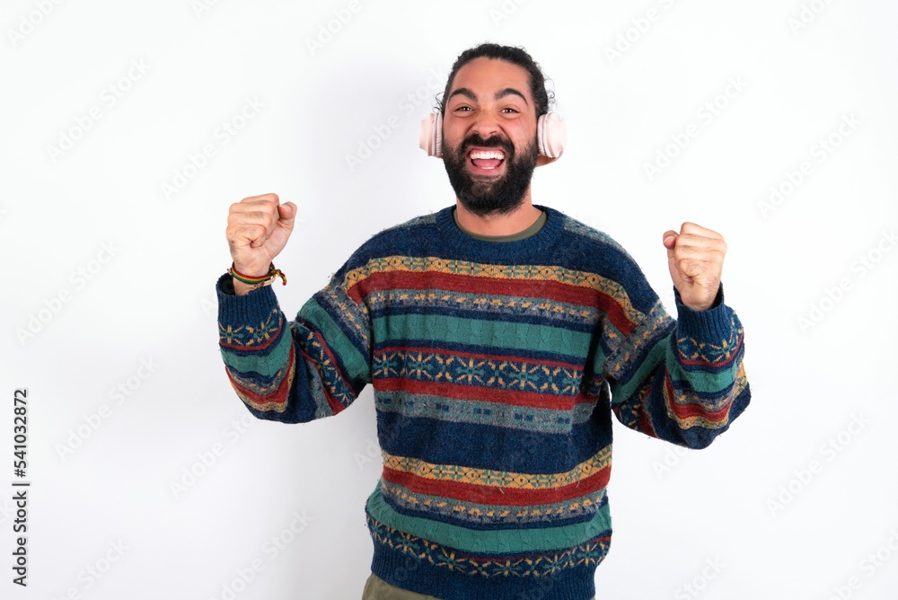 Naklejka premium Emotional Caucasian man with beard wearing sweater exclaims loudly feels like winner raises clenched fists keeps mouth opened wears stereo headphones on ears makes yes gesture, listens favorite music