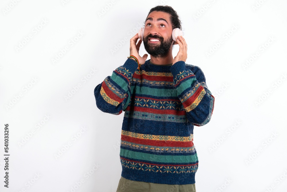 Joyful Caucasian man with beard wearing sweater over white background sings song keeps hand near mouth as if microphone listens favorite playlist via headphones Joyful Caucasian man with beard wearing sweater over white background sings song keeps hand near mouth as if microphone listens favorite playlist via headphones
