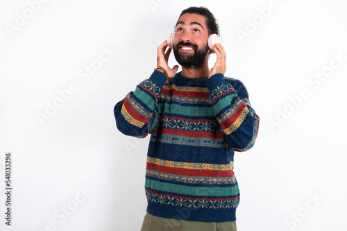 Joyful Caucasian man with beard wearing sweater over white background sings song keeps hand near mouth as if microphone listens favorite playlist via headphones