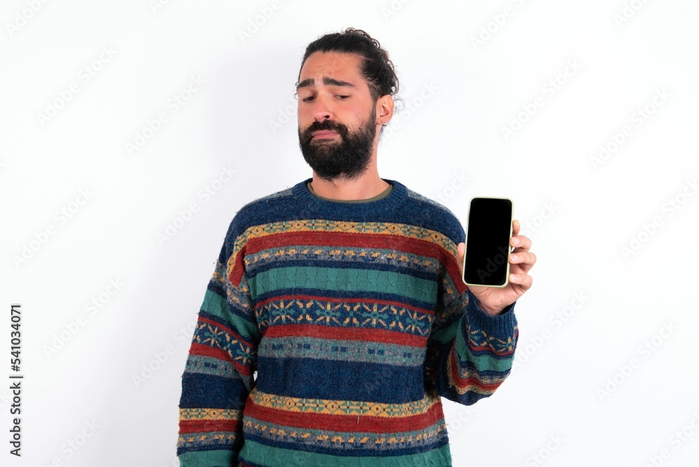 Photo of interested Caucasian man with beard wearing sweater over white background hold telephone look side empty space screen Photo of interested Caucasian man with beard wearing sweater over white background hold telephone look side empty space screen
