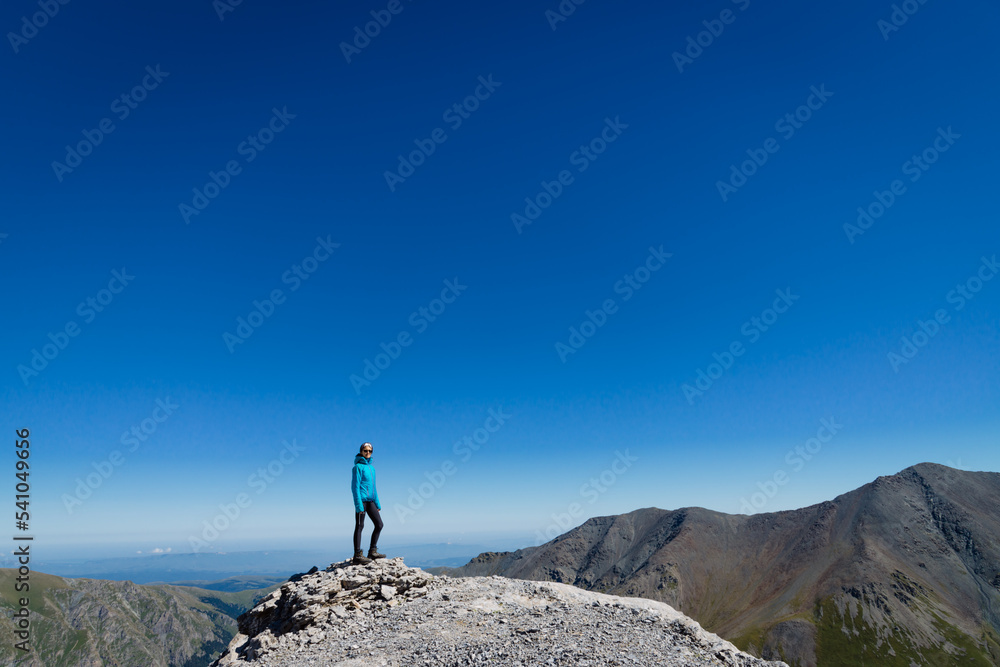 A woman stands in the mountains on top against the blue sky