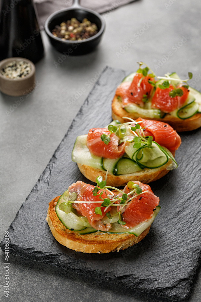 Healthy toasts with salmon, sliced cucumbers and microgreens . Bruschetta, snack, appetizers. Sandwiches on black stone board and grey background.