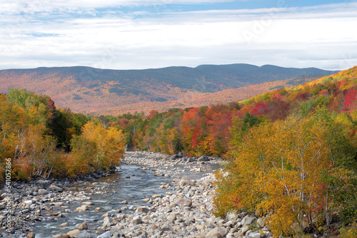 Scenic view of distant mountains in White Mountain National Forest and vibrant fall foliage along banks of rocky Pemigewasset River in New Hampshire.