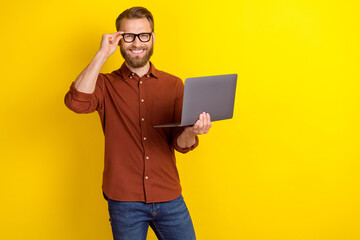 Photo of satisfied positive glad man blond beard dressed burgundy shirt hold laptop touch glasses isolated on yellow color background