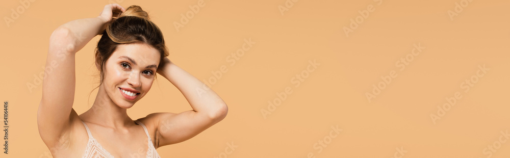 Obraz premium pretty and happy woman in bra looking at camera and fixing hair isolated on beige, banner