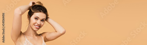 pretty and happy woman in bra looking at camera and fixing hair isolated on beige, banner