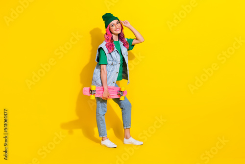 Full length photo of pretty cheerful girl dressed denim vest hand eyewear riding longboard isolated yellow color background