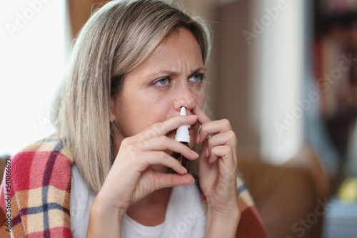 Ill woman sitting on sofa wrapped in blanket using nose drops