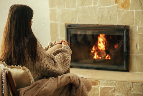Stylish woman in cozy sweater warming up in chair at fireplace in rustic room. Heating house in winter with wood burning stove. Young female sitting and relaxing at fireplace in farmhouse