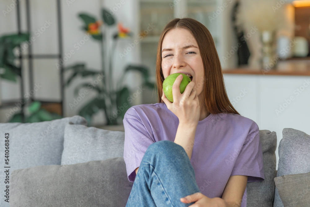 Healthy lifestyle concept. Beautiful young Caucasian woman biting fresh green apple sitting on the couch at home, looking at the camera Healthy lifestyle concept. Beautiful young Caucasian woman biting fresh green apple sitting on the couch at home, looking at the camera