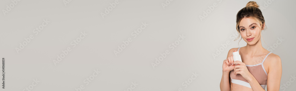 pretty woman in sports top holding container of vitamins and smiling at camera isolated on grey, banner pretty woman in sports top holding container of vitamins and smiling at camera isolated on grey, banner