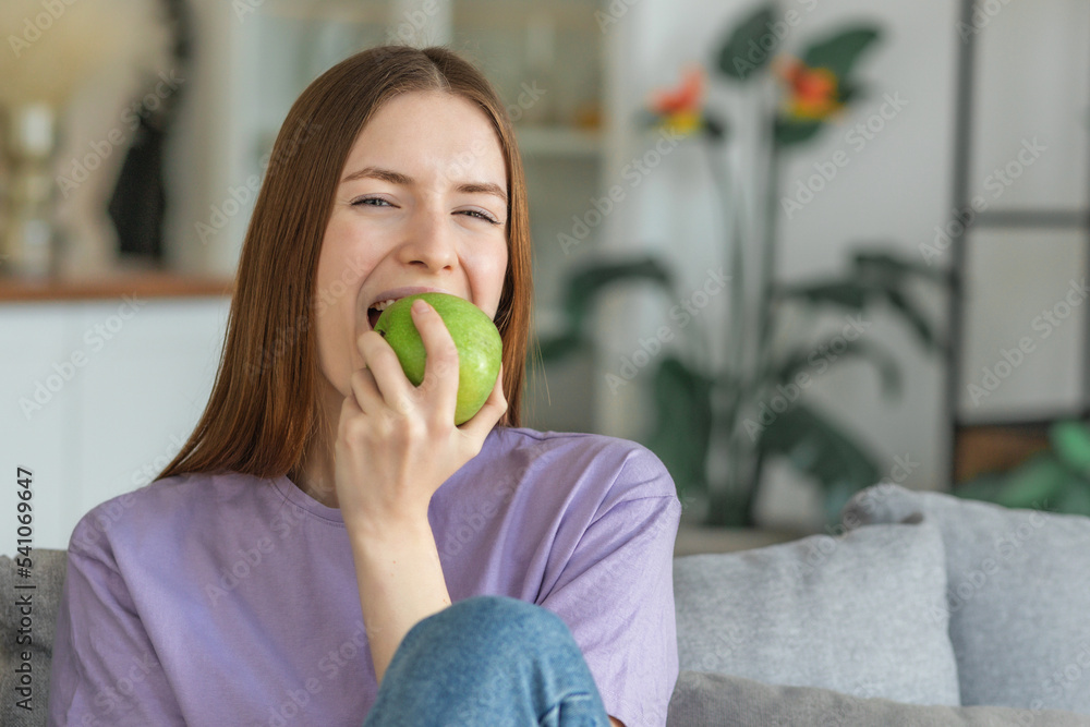 Healthy lifestyle concept. Beautiful young Caucasian woman biting fresh green apple sitting on the couch at home, looking at the camera Healthy lifestyle concept. Beautiful young Caucasian woman biting fresh green apple sitting on the couch at home, looking at the camera